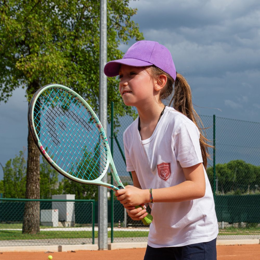 Bambina con cappellino e racchetta durante un corso di tennis su campo in terra rossa all'Adriano Panatta Racquet Club di Treviso