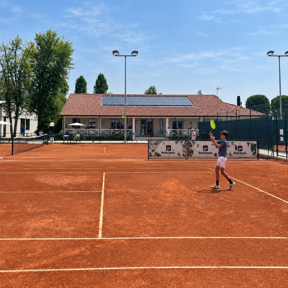 Giovane tennista su un campo in terra rossa all'aperto dell'Adriano Panatta Racquet Club a Treviso, con la club house sullo sfondo