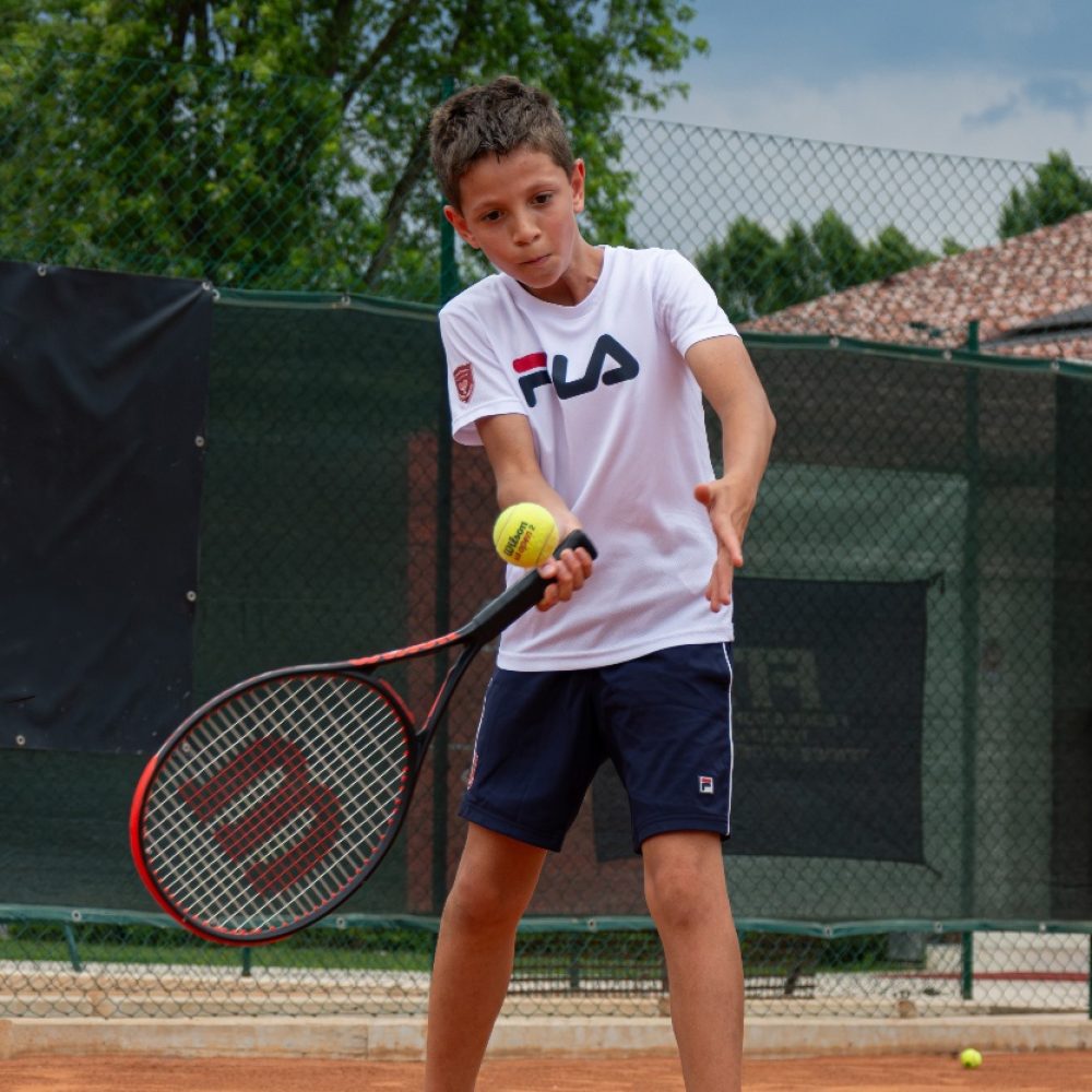 Bambino con maglia bianca che colpisce la palla da vicino con concentrazione durante il corso di tennis all'Adriano Panatta Racquet Club di Treviso