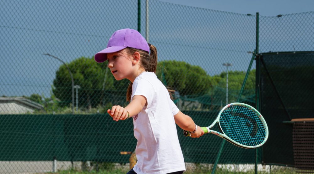 Bambina con cappellino viola e racchetta junior durante una lezione di mini tennis all'Adriano Panatta Racquet Club di Treviso