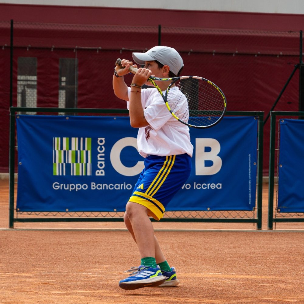Bambino con cappellino grigio che chiude il movimento del colpo con la racchetta sopra la spalla al corso di tennis dell'Adriano Panatta Racquet Club di Treviso
