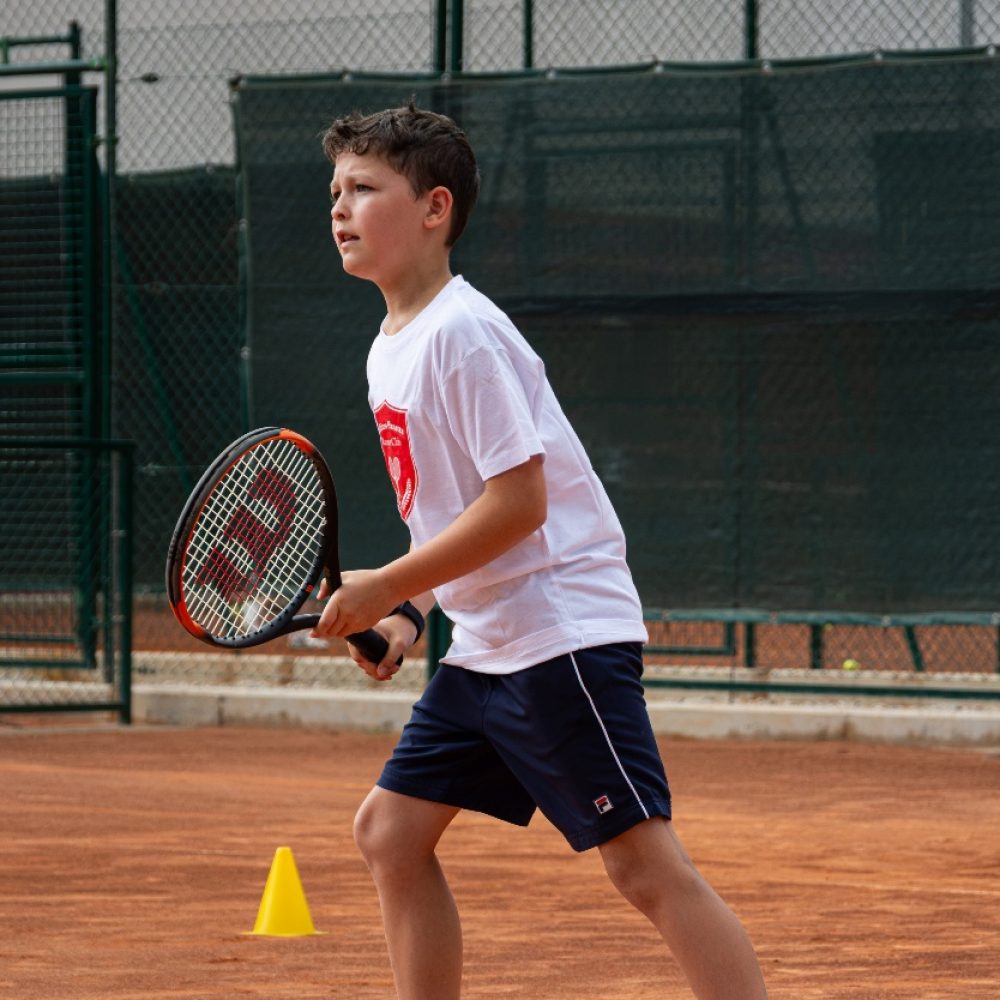 Bambino con maglia del club in posizione di attesa accanto a un cono durante il corso di tennis all'Adriano Panatta Racquet Club di Treviso