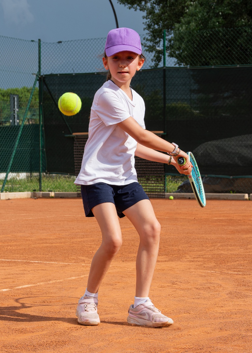 Bambina con cappellino viola che colpisce di rovescio a due mani su campo in terra rossa durante il corso di mini tennis all'Adriano Panatta Racquet Club di Treviso