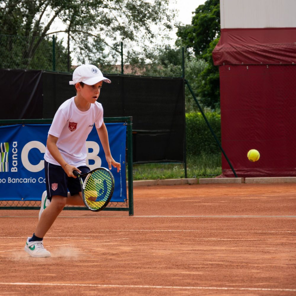 Bambino con cappellino bianco che corre verso la palla su campo in terra rossa durante l'allenamento della scuola tennis FITP a Treviso
